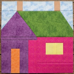 May include: A square fabric quilt block featuring a house design. The house has a purple wall, a green roof, a bright pink side wall, a yellow window, and an orange door. The background is light blue with clouds and two tan chimneys.