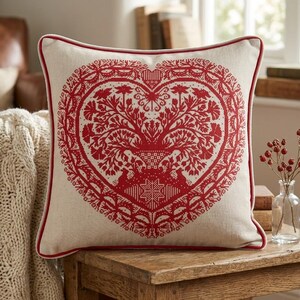 Op de afbeelding: Vierkant kussen met een crèmekleurige achtergrond en een rood hartvormig ontwerp. Het hart heeft een boom- en bloemmotief met een rode rand. Het kussen ligt op een houten tafel.