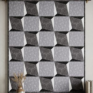 May include: A rectangular wall hanging quilt featuring a repeating geometric pattern of squares and triangles in shades of black, charcoal gray, and light gray. The fabric displays alternating damask and swirl motifs, suspended from a wooden rod.