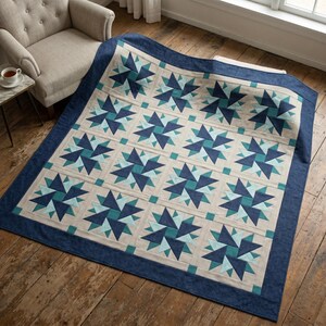 May include: A large, handmade quilt with a geometric star pattern in shades of blue and teal on a neutral background. The quilt has a dark blue border and is displayed on a wooden floor. The quilt is folded over a chair.
