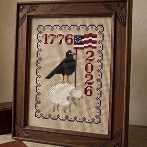 May include: Framed cross-stitch artwork featuring a black bird holding a flag with red and white stripes and blue stars, standing on a sheep. The text "1776" and "2020" are displayed. The frame is made of dark wood with carved corner details.