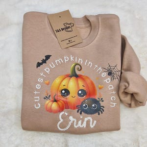 May include: A tan-coloured sweatshirt with a Halloween design. The graphic includes a cartoon pumpkin, spider, and the text "cutest pumpkin in the patch" and a name. A T&A Designs tag is attached.