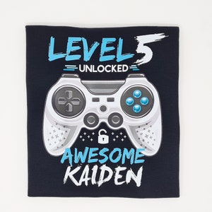 May include: Black t-shirt featuring a white video game controller graphic. The text "Level 5 Unlocked" and "Awesome Kaiden" is printed on the shirt.