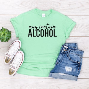 May include: A light green t-shirt with the text "may contain ALCOHOL" in black lettering. The shirt is laid flat on a white wooden surface with a pair of white trainers, a small potted succulent, and a pair of blue denim shorts.