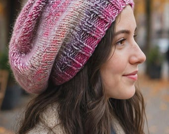 May include: A slouchy knit beanie featuring a variegated yarn pattern in shades of pink, purple, and cream. The hat is constructed with a thick, ribbed brim and a soft, textured stitch pattern that creates a relaxed, rounded silhouette.