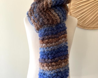 Puede incluir: Bufanda gruesa tejida a mano en tonos azules, marrones y beige, exhibida en un maniquí. La bufanda tiene una apariencia texturizada, con los colores mezclándose para crear un patrón único. El fondo es de color neutro.