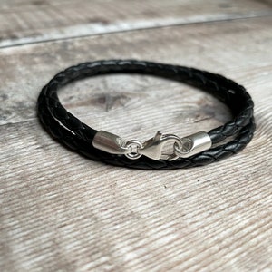May include: A black braided leather bracelet with a silver-colored clasp. The bracelet is wrapped around itself, showcasing the woven texture of the leather. The clasp is a lobster claw style.