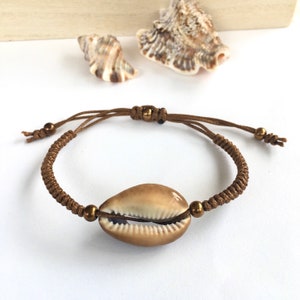 May include: A brown macrame bracelet featuring a natural cowrie shell centerpiece. The adjustable bracelet has bronze-colored beads and a sliding knot closure. The shell is a light tan color with a smooth, glossy finish. Seashells are in the background.