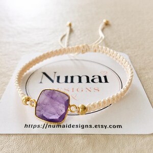 May include: A cream-colored macrame cord bracelet featuring a square, faceted purple amethyst stone set in a gold-toned bezel. The bracelet includes two small gold-toned beads flanking the stone and adjustable cord ends with gold-toned tips.