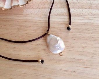 Pode incluir: Um colar com um pingente de pérola barroca branca em um cordão marrom e contas pretas. O colar é exibido em uma superfície de madeira.