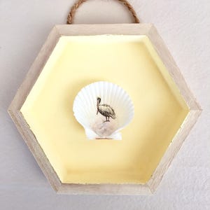 以下が含まれることがあります： 鳥のイラストが描かれた貝殻が特徴の、六角形の木製壁掛け。フレームは明るい黄色で、自然なロープのハンガーが付いています。貝殻が中心に配置され、海岸をテーマにしたデザインになっています。
