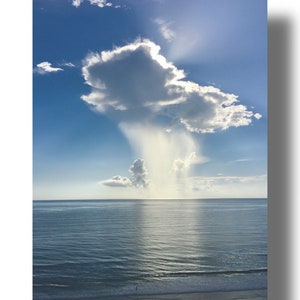 May include: A large, white cloud with a sunbeam shining through it, over a blue ocean with a sandy beach in the foreground.