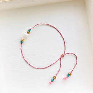 May include: A pink string bracelet with a white shell charm and gold and turquoise beads.