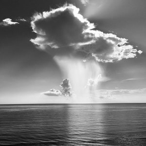 Op de afbeelding: Zwart-witfoto van een zeegezicht met een dramatische lucht. Een grote, pluizige wolk domineert het bovenste deel, met zonnestralen die erdoorheen breken. De oceaan strekt zich uit tot aan de horizon en weerspiegelt het licht.