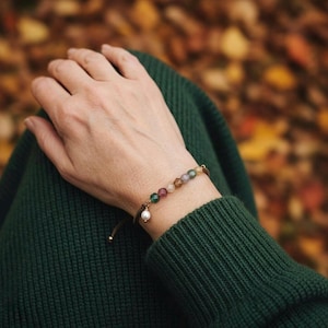 May include: A delicate bracelet featuring a series of small, multi-coloured faceted gemstone beads arranged in a row, accented by a single round white pearl charm dangling from a thin, dark brown cord with an adjustable sliding knot closure.
