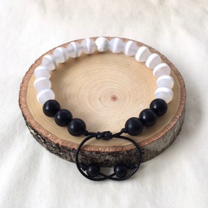 Op de afbeelding: Een witte en zwarte kralen armband met een lava kraal en een oneindigheids knoop sluiting.