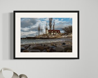 May include: A framed photograph of a lighthouse on a rocky shoreline under a cloudy sky. The lighthouse has a white tower and a red roof. The foreground features a beach with pebbles and the ocean. The photograph is displayed on a black table.