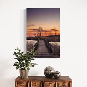 May include: A wooden bridge extends over a still body of water, leading to a small island with trees. The sky is a vibrant orange and purple, creating a beautiful sunset scene.