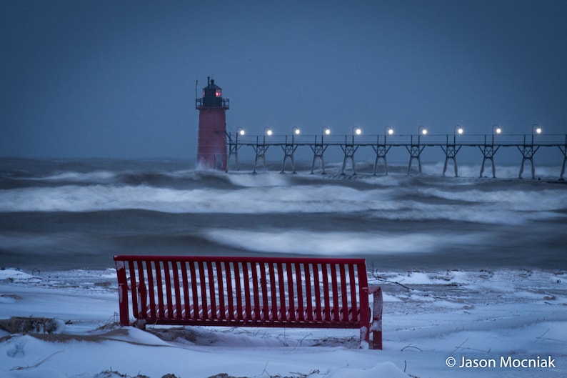 South Haven Lighthouse with a red bench and a snow covered beach, white capped waves of Lake Michigan. Evening/night with lights lit up on the lighthouse.