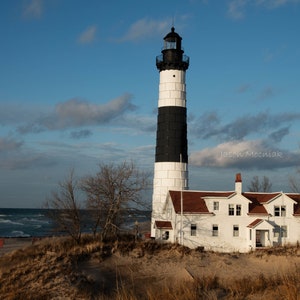 Puede incluir: Un faro blanco y negro con un techo rojo se alza sobre una playa de arena con un cielo azul y nubes blancas en el fondo. El faro es alto y delgado, con una base blanca y una parte superior negra. El techo es rojo y tiene una peque&ntilde;a ventana a cada lado. La playa es arenosa y tiene algunos &aacute;rboles en el fondo. El cielo es azul y tiene algunas nubes blancas.