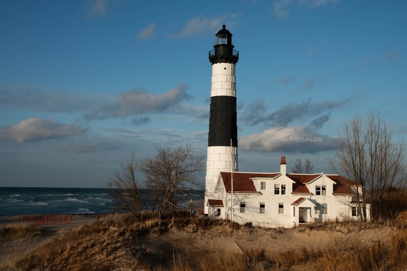 Puede incluir: Un faro blanco y negro con un techo rojo se alza sobre una playa de arena con un cielo azul y nubes blancas en el fondo. El faro es alto y delgado, con una base blanca y una parte superior negra. El techo es rojo y tiene una peque&ntilde;a ventana a cada lado. La playa es arenosa y tiene algunos &aacute;rboles en el fondo. El cielo es azul y tiene algunas nubes blancas.