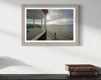 May include: A framed photograph of a lighthouse with a view of the sea. The lighthouse is made of brick and has a white roof. The sea is blue and calm. The sky is cloudy and grey.