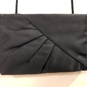 Könnte beinhalten: Eine schwarze Stoff-Clutch mit plissiertem Design und einem schwarzen Riemen.