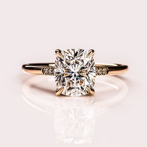 May include: A gold engagement ring with a large, square-cut diamond as the centerpiece. The band is thin and gold, with small accent diamonds on each side of the main stone. The ring is set against a soft, light background.