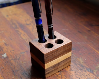 May include: A wooden pen holder with four slots, made of walnut and maple wood. Two pens, one blue and one red, are in the holder.