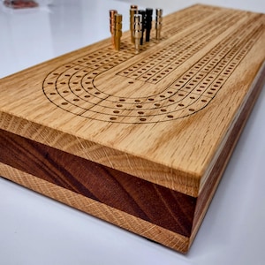May include: A rectangular cribbage board made of light-colored wood with a dark wood center stripe. The top surface features laser-engraved tracks and holes for scoring. Several metallic pegs in copper, silver, and black are inserted into the board.