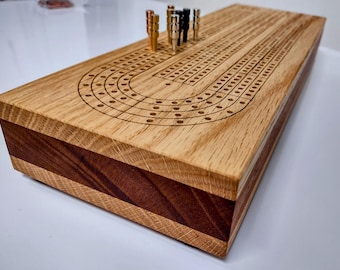 May include: A rectangular cribbage board made of light-colored wood with a dark wood center stripe. The top surface features laser-engraved tracks and holes for scoring. Several metallic pegs in copper, silver, and black are inserted into the board.