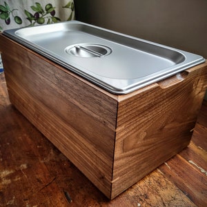 May include: A wooden countertop compost bin with a stainless steel lid. The bin has a natural wood finish and is made of solid wood.