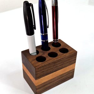 May include: A wooden pen holder with six circular openings, holding a black marker, a blue pen, and a red pen. The holder is made of dark brown wood with a lighter brown stripe. The pens are visible.