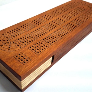 May include: A rectangular wooden cribbage board featuring a dark brown top surface with engraved black playing tracks and holes. The sides of the board are constructed from alternating layers of light and dark wood, creating a striped pattern.