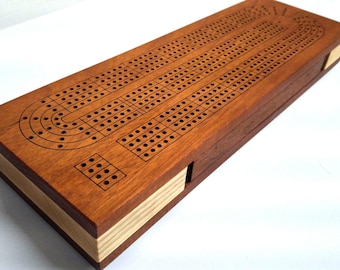May include: A rectangular wooden cribbage board featuring a dark brown top surface with engraved black playing tracks and holes. The sides of the board are constructed from alternating layers of light and dark wood, creating a striped pattern.