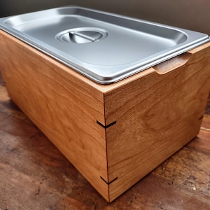 May include: A wooden box with a stainless steel lid. The box is made of cherry wood and has a natural finish. The lid has a small handle on the side.