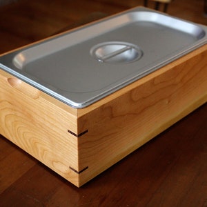 May include: A wooden box with a stainless steel lid. The box is made of light wood with a natural finish. The lid has a handle and a slightly raised edge.