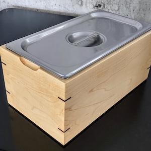May include: A wooden box with a stainless steel insert. The box is made of light wood with a natural finish. The insert is smooth and shiny.