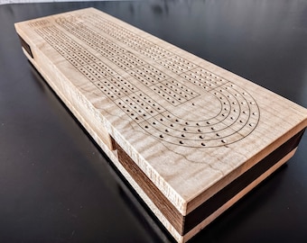 May include: A rectangular wooden cribbage board featuring a light-colored, wavy-grained top surface with laser-engraved tracks and holes for scoring. The sides of the board are constructed with a contrasting dark brown wood inlay layer.