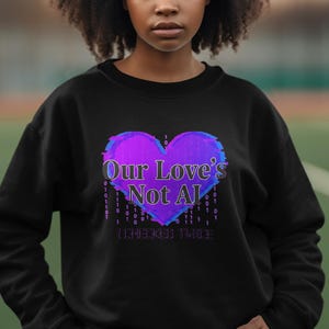 Pode incluir: Camisola preta com um gráfico de coração roxo e azul vibrante com o texto "Our Love's Not AI" e "I Remember Thee". O design inclui elementos de código binário. Uma parte de cima casual e confortável.