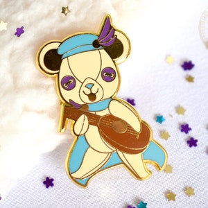 Könnte beinhalten: Goldfarbene Emaille-Anstecknadel mit einem weißen Bären, der einen blauen Hut mit einer violetten Feder trägt und eine braune Laute spielt. Der Bär hat violette Augen und einen blauen Schal. Die Anstecknadel ist von goldenem, violettem und blauem Glitzer umgeben.