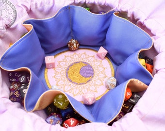 Puede incluir: Una bolsa de dados lila con interior azul y un diseño bordado en rosa. La bolsa contiene varios dados coloridos. El diseño bordado presenta una luna creciente y un patrón de sol. La bolsa está abierta, revelando los dados en su interior.