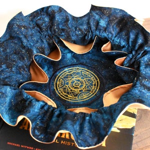 May include: A round, open-topped fabric container with a celestial design. The exterior is a dark blue with gold flecks, and the interior is tan. A gold embroidered symbol is centered on the bottom. The container is sitting on top of a book.
