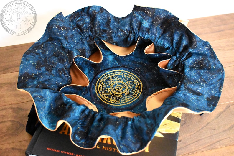 May include: A round, open-topped fabric container with a celestial design. The exterior is a dark blue with gold flecks, and the interior is tan. A gold embroidered symbol is centered on the bottom. The container is sitting on top of a book.