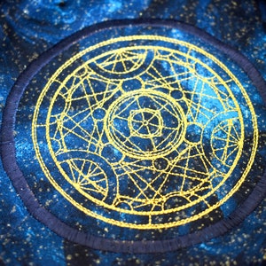 May include: A circular embroidered patch with a celestial design. The patch features a gold-colored geometric pattern on a dark blue background with a navy blue border. The background has a speckled, starry effect.