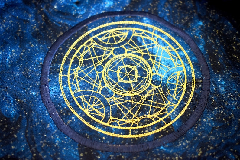 May include: A circular embroidered patch with a celestial design. The patch features a gold-colored geometric pattern on a dark blue background with a navy blue border. The background has a speckled, starry effect.