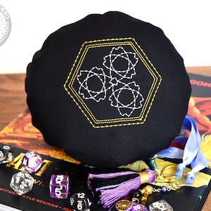 May include: Black dice bag with a gold and white geometric design embroidered on the front. The bag is sitting on a book with the word "DRAGONS" visible on the cover. There are several dice scattered around the bag.