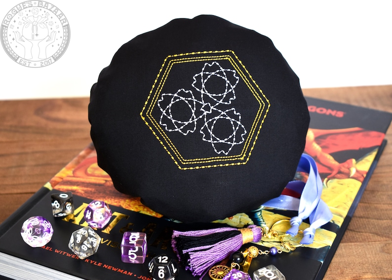 May include: Black dice bag with a gold and white geometric design embroidered on the front. The bag is sitting on a book with the word "DRAGONS" visible on the cover. There are several dice scattered around the bag.