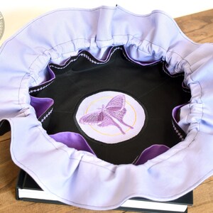 May include: A lavender fabric bowl with a black lining and a purple embroidered moth design. The bowl is sitting on a black book.