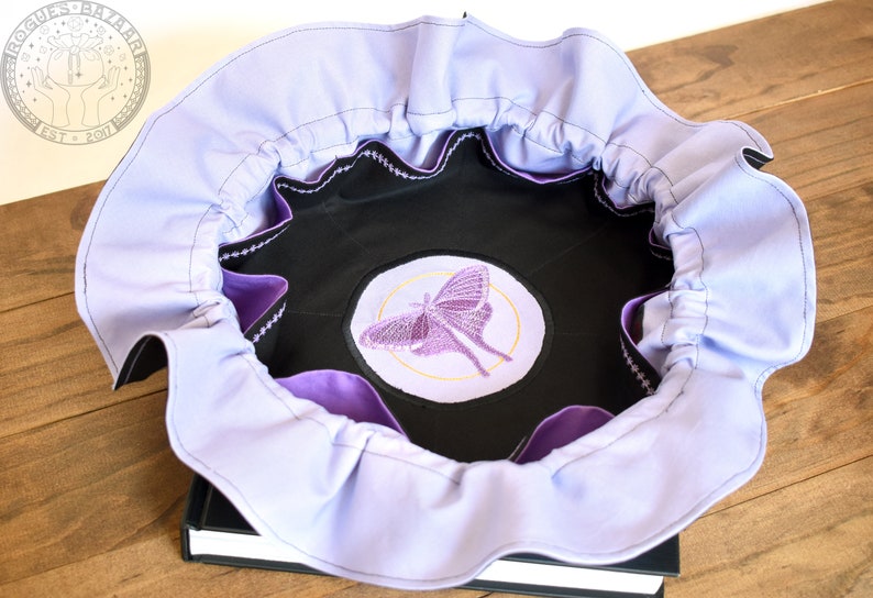 May include: A lavender fabric bowl with a black lining and a purple embroidered moth design. The bowl is sitting on a black book.
